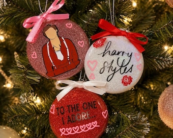 May include: Three round glittery Christmas baubles hanging on a tree. One is pink with a cartoon illustration of a person in red braces, one is white with the text "harry styles" and red roses, and one is red with the text "TO THE ONE I ADORE".