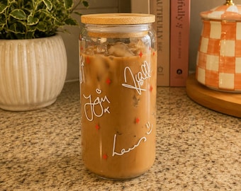 May include: A clear glass tumbler filled with iced coffee, featuring a bamboo lid and white script text that reads "all you need is love" along with small red four-pointed star accents printed on the exterior surface of the glass.
