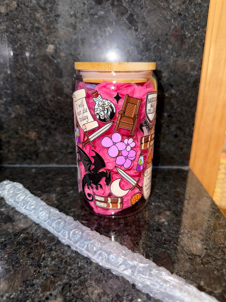May include: A clear glass tumbler with a pink background and various illustrations, including a dragon, a sword, a crown, a potion bottle, and a book. The tumbler has a bamboo lid and the text "I will not die today" and "Based God Collector" are visible on the tumbler.