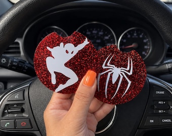 May include: Two circular car cup holder coasters featuring a red glitter finish. One coaster displays a white silhouette of a superhero in a dynamic pose, while the other features a white spider emblem. Both coasters have a small notch on the edge.