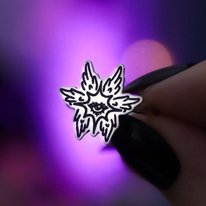 May include: A silver pin with a black and white design of an eye surrounded by wings. The pin is being held by a hand with black nail polish.