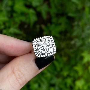 May include: A silver ring with a square face featuring a detailed black and white illustration of two frogs. The ring has a minimalist design, perfect for everyday wear. The ring is held up against a green leafy background.