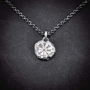 May include: A silver pendant necklace with a circular pendant etched with a Viking compass design. The pendant is suspended from a fine chain. The background is dark grey.
