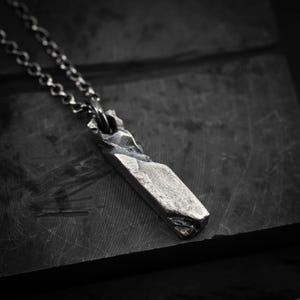 May include: A silver pendant necklace featuring a rectangular, textured pendant. The pendant is suspended from a chain. The necklace is shown on a dark, textured surface.