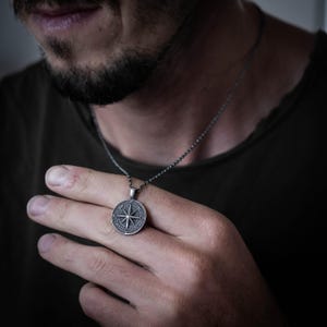 May include: A silver pendant necklace with a compass design. The round pendant displays a detailed compass rose and hangs from a fine chain. The necklace is set against a dark backdrop.