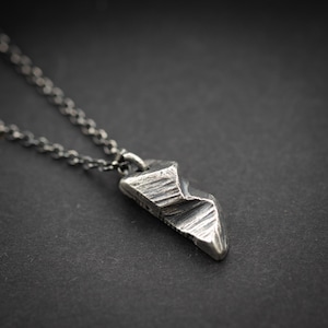 May include: A silver pendant necklace featuring a textured, geometric design. The pendant, resembling a mountain or arrowhead, hangs from a fine chain. The necklace is set against a dark backdrop, accentuating the pendant's details and the chain's links.