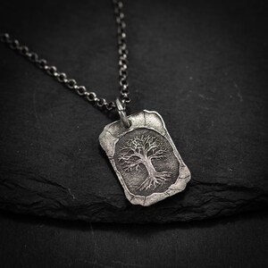 May include: A silver-toned pendant necklace featuring a detailed tree design within a rectangular frame. The pendant hangs from a matching chain. The tree's branches and roots are intricately rendered, creating a textured, nature-inspired aesthetic. The necklace is displayed against a dark background.