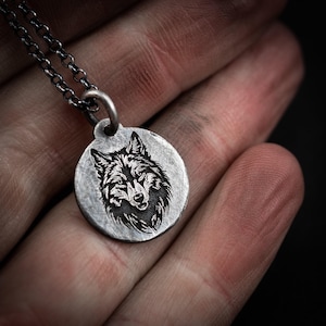 Può includere: Una collana con pendente in argento con un design dettagliato a testa di lupo. Il pendente rotondo è attaccato a una delicata catena. I lineamenti del lupo sono incisi in modo intricato, mostrando la sua pelliccia e la sua espressione. È un gioiello.
