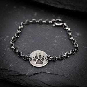 May include: A silver-toned bracelet featuring a circular charm with a paw print design. The charm is connected to a chain-link bracelet with a lobster clasp. The bracelet is displayed on a dark, textured surface.