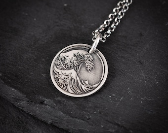 May include: A silver-colored pendant necklace featuring a detailed wave design within a circular frame. The pendant hangs from a delicate chain, set against a dark, textured background. The wave design is intricately carved, showcasing the cresting water.