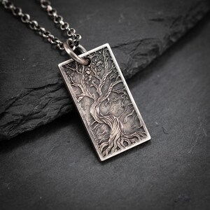 Puede incluir: Un colgante rectangular plateado con un diseño detallado de árbol de la vida. El colgante cuelga de una cadena plateada. El árbol presenta un tronco retorcido, ramas y hojas. El colgante mide aproximadamente 3,8 cm de largo.