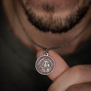 May include: A silver pendant necklace with a detailed circular medallion. The medallion shows a winged figure holding a shield and sword. The necklace chain is a dark silver colour.
