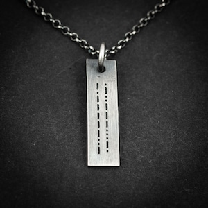 Pode incluir: Um colar com pingente prateado com uma etiqueta retangular com pontos e traços gravados, provavelmente código Morse. O pingente pende de uma corrente escura e texturizada sobre um fundo preto. Um acessório minimalista.