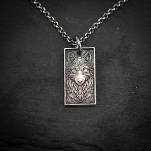 Op de afbeelding: Een zilverkleurige rechthoekige hangerketting met een gedetailleerde wolfskop. Het gezicht van de wolf is ingewikkeld gesneden, met vacht en gelaatstrekken. De hanger hangt aan een bijpassende zilveren ketting.