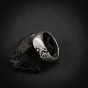 May include: A silver ring with a textured design, resting on a dark, rough surface. The ring has a smooth, curved band and a detailed, possibly carved, central element. The background is a solid black.