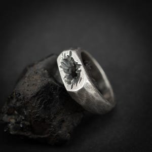May include: A silver ring with a textured, rectangular face featuring a carved design. The ring is set on a dark, rough stone against a black background. The ring's band has a brushed metal finish.
