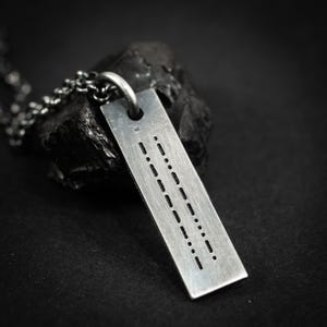 Può includere: Una collana con pendente rettangolare argentato con un design a codice Morse. Il pendente è sospeso a una catena e presenta una serie di punti e trattini. La collana è esposta su una superficie scura e strutturata.