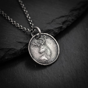 Op de afbeelding: Een zilverkleurige ketting met een gedetailleerd reliëf van een hert. De ronde hanger hangt aan een fijne ketting. Het hert is in profiel afgebeeld, met een imposant gewei. De ketting ligt op een donkere, getextureerde ondergrond.