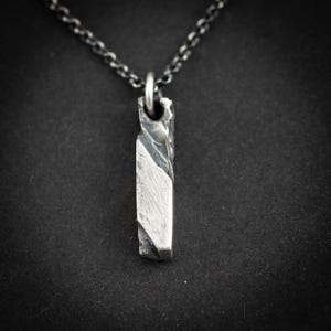 Könnte beinhalten: Eine silberne Halskette mit einem rechteckigen, strukturierten Anhänger. Der Anhänger hat ein raues, asymmetrisches Design mit einem diagonalen, glatten Abschnitt. Die Kette ist dunkel und hat Glieder. Der Hintergrund ist schwarz.