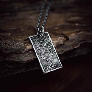 May include: A silver-colored pendant necklace featuring a detailed tree design within a rectangular frame. The tree's branches and roots are intricately carved, creating a textured appearance. The pendant hangs from a delicate chain, showcasing a nature-inspired theme.