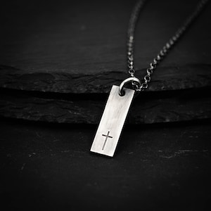 May include: A silver pendant necklace with a rectangular tag and a cross etched on the front. The pendant is suspended from a fine chain. The dark background accentuates the necklace's design.
