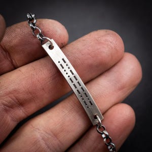 May include: A silver bracelet with a rectangular bar etched with Morse code. The bar is connected to a chain-link bracelet. The bracelet is held in a hand against a dark background.