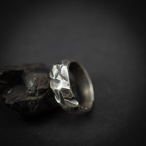 Puede incluir: Un anillo de plata texturizado con un diseño geométrico. La superficie del anillo presenta facetas angulares, dándole un aspecto robusto. El anillo se muestra sobre un fondo oscuro. Un accesorio moderno, con un diámetro aproximado de 2 cm.