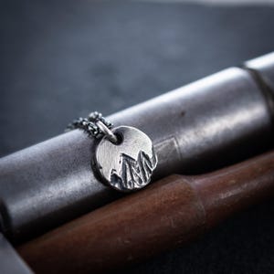 Puede incluir: Un collar con colgante plateado con un diseño de cordillera. El colgante redondo está sujeto a una cadena. El collar se exhibe sobre un objeto metálico con un mango de madera.
