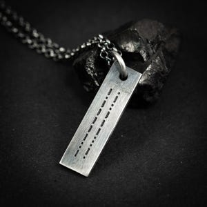 Puede incluir: Un collar con colgante plateado con una placa rectangular con un patrón de código Morse. El colgante cuelga de una cadena plateada. El fondo es una superficie oscura y texturizada.
