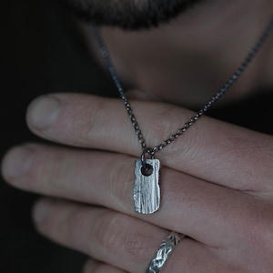 Puede incluir: Collar con colgante plateado de forma rectangular y texturizada. El colgante cuelga de una fina cadena. El collar se sostiene en una mano, con un anillo plateado visible. El fondo está borroso.