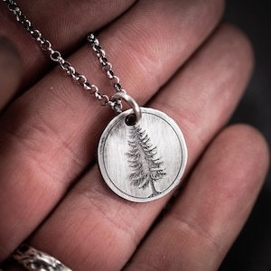 Può includere: Una collana con pendente in argento con un ciondolo circolare inciso con un design dettagliato di pino. Il pendente è sospeso a una sottile catena d'argento. La collana è mostrata su uno sfondo sfocato.