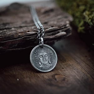 Puede incluir: Un collar con colgante plateado con una imagen detallada de Buda. El colgante circular cuelga de una fina cadena. El collar se exhibe sobre una superficie de madera, con un fondo borroso.