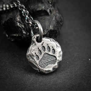 Puede incluir: Un collar con colgante plateado con una huella de pata de oso. El colgante redondo tiene una superficie texturizada y rugosa, y la huella de la pata está detallada con acentos oscuros. La cadena también es plateada.