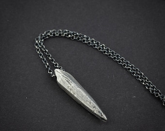 May include: A silver pendant necklace featuring a textured, elongated arrowhead design. The pendant hangs from a delicate chain. The necklace is set against a dark background, accentuating the metallic sheen.