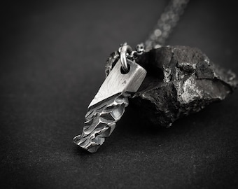 May include: A silver pendant necklace with a textured, geometric design. The pendant has a rectangular top and a jagged, rough-hewn lower edge. The necklace chain is silver and rests on a dark surface.