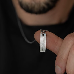 Peut inclure: Un collier avec un pendentif rectangulaire argenté orné d'une petite croix. Le pendentif est suspendu à une chaîne délicate. Le collier est présenté sur un fond sombre.