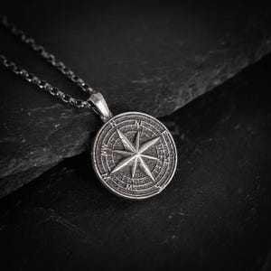 May include: A silver-colored compass pendant necklace. The pendant features a detailed compass rose design with directional markers. The necklace chain is also silver and has a link design. The pendant is round and has a textured surface.
