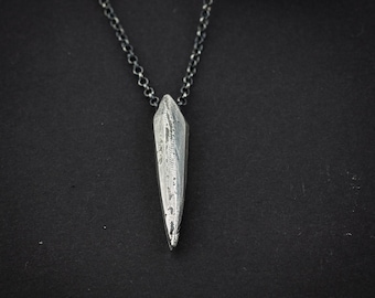 May include: A silver pendant necklace with a textured, elongated crystal-shaped charm. The charm hangs from a delicate chain against a dark background. The pendant has a pointed tip and detailed surface.