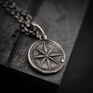 Könnte beinhalten: Eine silberfarbene Halskette mit Anhänger, die ein Kompassrosen-Design aufweist. Der runde Anhänger hat eine strukturierte Oberfläche mit einem erhabenen Sternmuster und hängt an einer Kette. Die Halskette wird auf einer dunklen, strukturierten Oberfläche präsentiert.