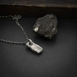 Op de afbeelding: Een zilveren ketting met een rechthoekige hanger. De hanger heeft een ruw, getextureerd oppervlak en een kleine ring aan de bovenkant.