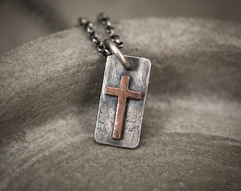 May include: A silver-tone pendant necklace featuring a rectangular tag with a copper cross. The cross is centered on the tag, and the necklace has a dark chain. The pendant is displayed on a textured, gray surface.