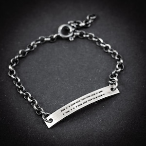 Op de afbeelding: Een zilverkleurige armband met een schakelkettingontwerp. De armband heeft een rechthoekige staaf met morsecode-symbolen. De armband wordt vastgezet met een sluiting. De sieraden worden op een donkere achtergrond getoond.