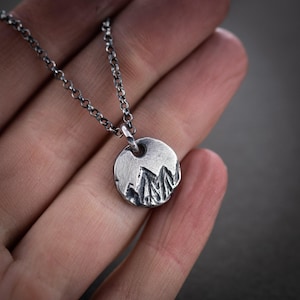 Peut inclure: Un collier pendentif argenté avec un charm circulaire représentant une chaîne de montagnes. Le charm est suspendu à une fine chaîne. Le collier est tenu dans une main, mettant en valeur le design et la texture du pendentif.