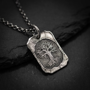 Puede incluir: Un collar con colgante plateado con un diseño detallado de árbol dentro de un marco rectangular. El colgante cuelga de una cadena a juego. Las ramas y raíces del árbol están intrincadamente representadas, creando un accesorio texturizado inspirado en la naturaleza.
