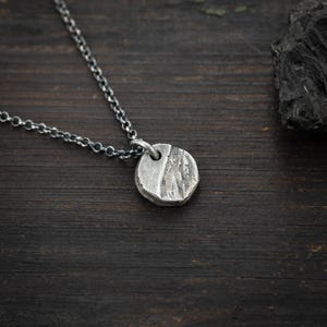 Op de afbeelding: Een zilveren hangerketting met een getextureerde, cirkelvormige hanger. De hanger is bevestigd aan een fijne ketting. De ketting wordt gepresenteerd op een donker houten oppervlak.