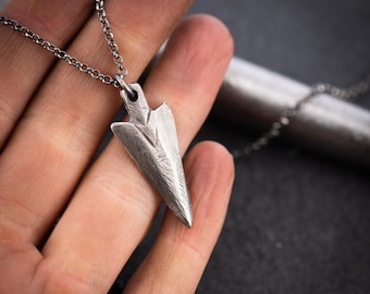 May include: A silver arrowhead pendant necklace. The pendant, with etched details, is shaped like an arrowhead and hangs from a delicate chain. The necklace is held in a hand against a dark background.
