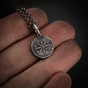 Könnte beinhalten: Halskette mit Kompass-Anhänger aus Silber. Der runde Anhänger zeigt ein detailliertes Kompassrosen-Design. Die Halskette hat eine Gliederkette. Anhänger und Kette werden in einer Hand vor dunklem Hintergrund gehalten.