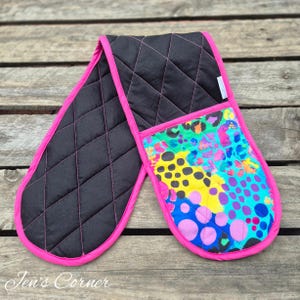 May include: A double oven glove with a black quilted exterior, pink trim, and stitching. One side has a colourful abstract pattern with blue, yellow, and pink spots. The other side is solid black.