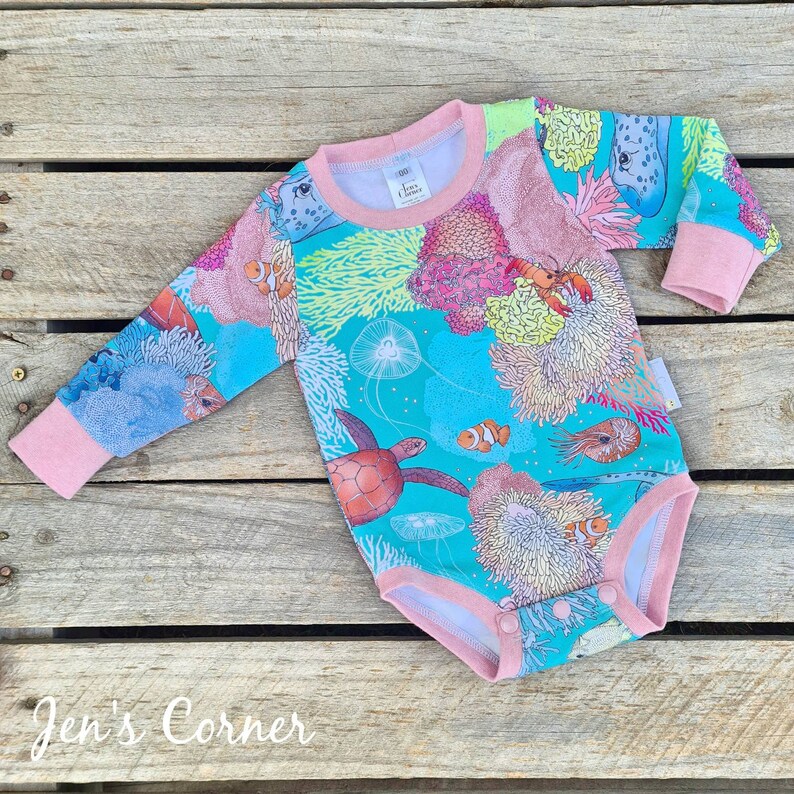 May include: A turquoise long-sleeved baby onesie with a colorful coral reef print. The onesie features a variety of sea creatures, including fish, sea turtles, and jellyfish. The onesie has pink trim and snaps at the bottom.