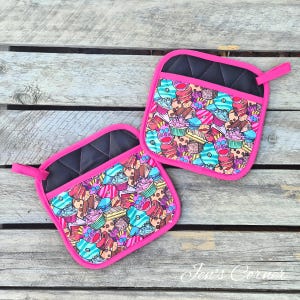 May include: Two square oven mitts with a black quilted top and a bright pink border. The main pattern features colourful cupcakes, doughnuts, and other sweets. Each mitt has a pink loop for hanging.